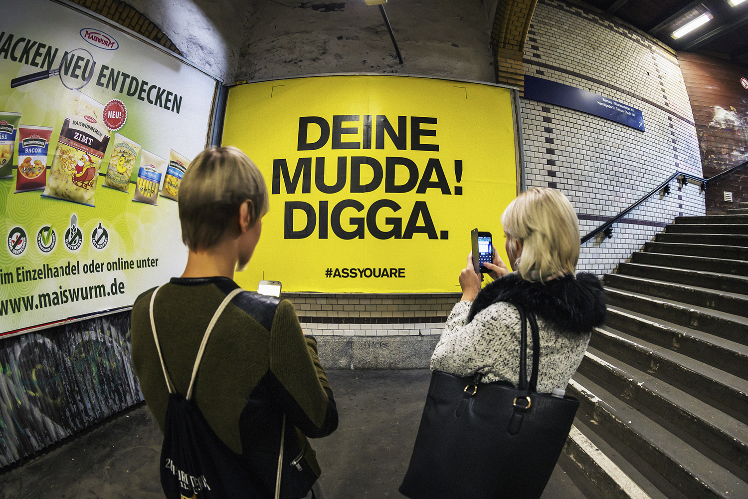 Plakatwand mit gelbem Plakat in einer Bahnunterführung, die von zwei Personen fotografiert wird
